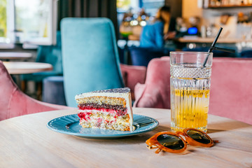 Beautiful glass of yellow passionfruit lemonade and piece of poppy cake with brown sunglasses on the wooden table. Flatlay style, spotted on the city cafe. Summer brunch situation.