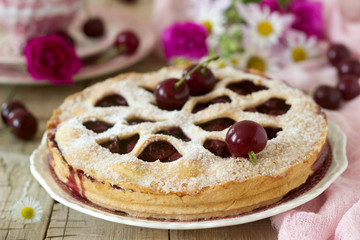 A cherry pie decorated with heart-shaped ornamentation with a cup of tea, a bouquet of flowers and ripe cherry berries.