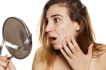 young shocked woman with problematic skin and without makeup looking her self in the mirror on a...