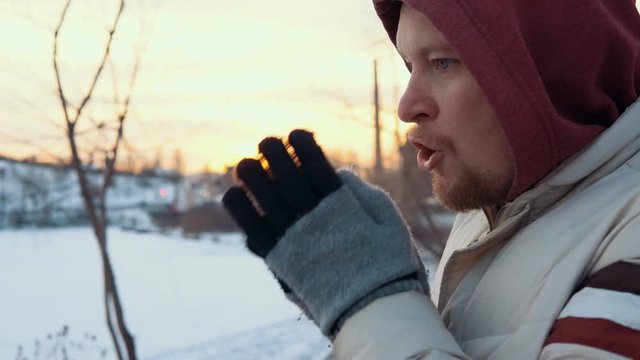 Portrait of a young red-bearded man standing frozen against a city landscape. The man puts his hands in gloves to his mouth and exhales warm air, tries to warm up.