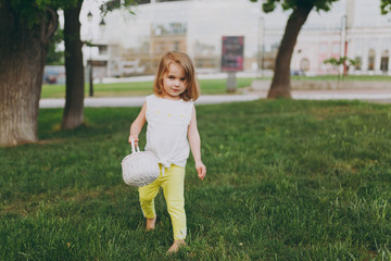 Fototapeta premium Pretty little cute child baby girl with basket walking, play and have fun on green grass lawn in park. Little kid daughter in yellow clothes. Mother's Day, love family, parenthood, childhood concept.