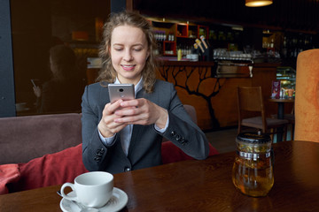 Young business woman writing a message on smart phone and smiling at cafe