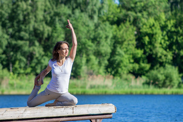 Fototapeta na wymiar Young caucasian woman doing yoga asanas outdoors on a wooden pier by the river or lake, sunny summer morning, wearing no makleup and sports clothes