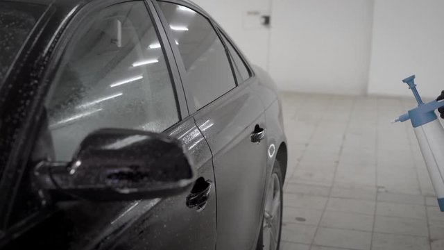 washerman is spraying water by nebulizer on a black car body in a car wash box, close-up of hands