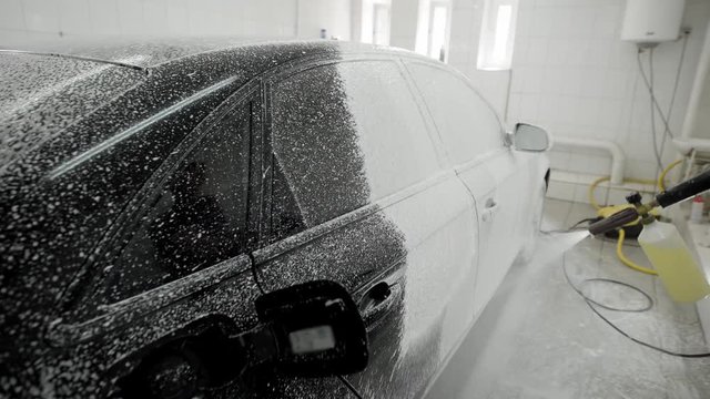 worker is covering car body by soap foam for washing in a garage, using hose for spraying, car washing