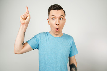 Young thinking guy dressed in a blue t-shirt shows finger up with wide eyes and opened mouth on a light background.