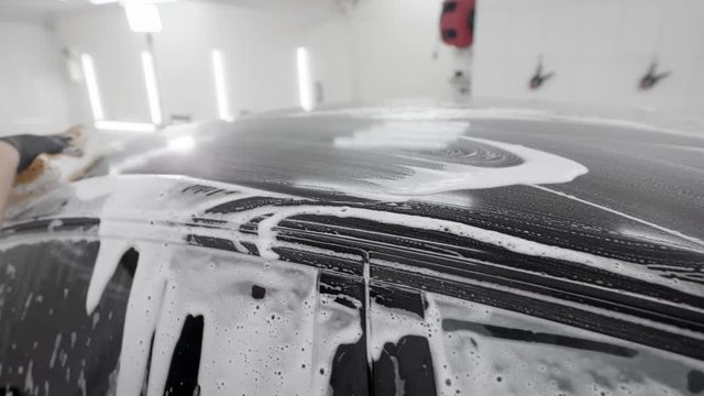 washerman is rubbing surface of black automobile by sponge with soap foam in car washing, wearing rubber gloves