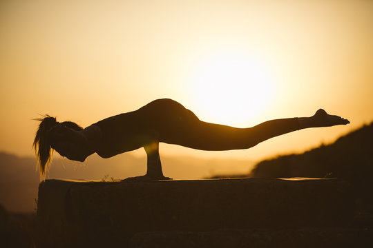 Young Woman Is Practicing Yoga On The Mountain At Sunset. Silhouette Of Young Woman Practicing Yoga Outdoor.