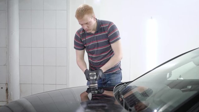 redhead male worker is processing surface of car by polishing machine in a car-service, covering by protective layer
