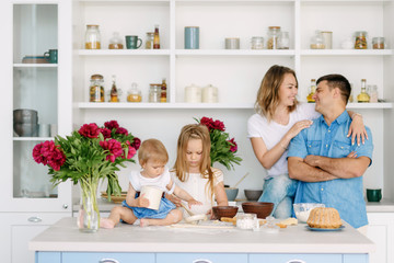 Young parents prepare a delicious and healthy breakfast in a large bright kitchen with their two lovely children. Dad kisses and hugs mother, children are happy