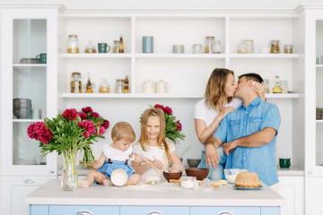 Young parents prepare a delicious and healthy breakfast in a large bright kitchen with their two lovely children. Dad kisses and hugs mother, children are happy