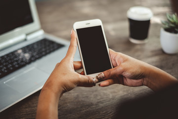 Close up business woman's hand holds smart phone with black isolated screen over background of laptop, coffee on wooden background