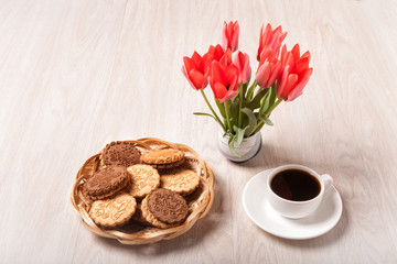 Cookies in a wicker basket, white Cup with coffee on a saucer and a bouquet of tulips on a light textured wooden surface