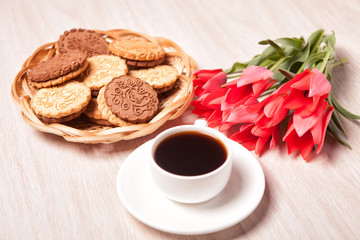 Cookies in a wicker basket, white Cup with coffee on a saucer and a bouquet of tulips on a light textured wooden surface