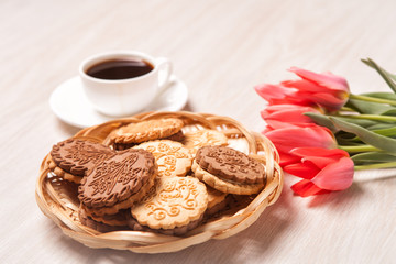 Cookies in a wicker basket, white Cup with coffee on a saucer and a bouquet of tulips on a light textured wooden surface