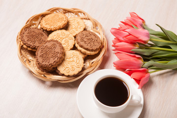 Cookies in a wicker basket, white Cup with coffee on a saucer and a bouquet of tulips on a light textured wooden surface