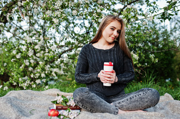 Young brunette girl at jeans sitting on plaid against spring blossom tree and holding tea thermos at hands.