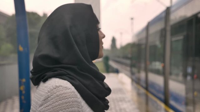 Young muslim woman in hijab is watching how train is coming under raining, religion concept, transport concept, weather concept