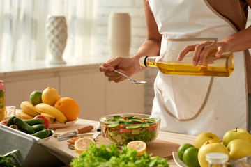 Female food blogger wearing apron pouring olive oil into vegetable salad while standing at modern...