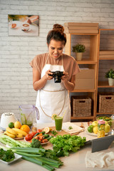 Attractive middle-aged food blogger wearing apron taking picture of healthy smoothie while standing at modern kitchen