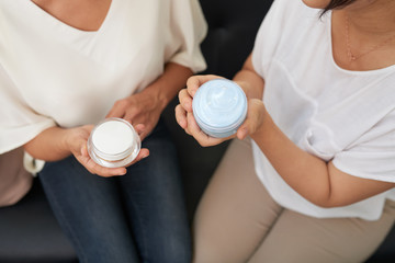 Two jars with facial rejuvenation cream in hands of two young women during discussion of their effectiveness