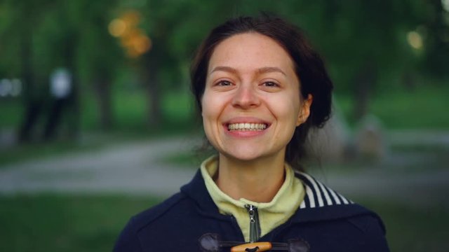 Slow motion portrait of charming young woman wearing casual clothes standing outside in park and smiling looking at camera. People, nature and healthy lifestyle concept.