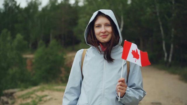 Slow motion portrait of female traveller pretty girl holding Canadian flag, smiling and looking at camera with beautiful natural landscape in background.