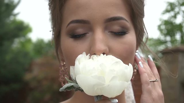 Close-up portrait of the charming bride with natural make-up smelling the white peony with closed eyes.