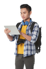 Concentrated young traveler with backpack standing against white background and studying map on digital tablet, portrait shot