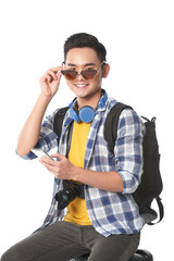 Cheerful Asian tourist sitting on suitcase and peering over sunglasses while posing for photography, he studying map on smartphone, isolated on white background