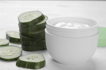 Face cream in a white jar with cucumber extract next to a set of fresh cucumbers on a white wooden background