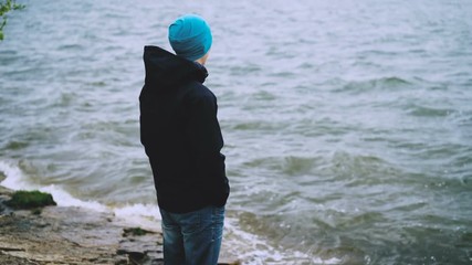 Slow motion portrait of thoughtful guy in hat, jacket and jeans standing on the sea coast and watching water waves enjoying nature. Healthy lifestye and tourism concept.