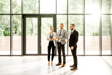 Group of a young strictly dressed business people standing together with digital tablet at the modern office hall indoors