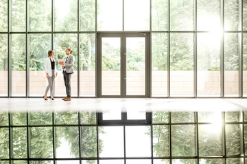 Fototapeta na wymiar Business couple standing together at the modern office hall with window on the background overlooking on the park. View directly with reflection from the table
