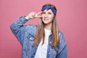 Stylish beautiful female hippie wears trendy oversized denim jacket, blue headband and white t shirt, smiling gently and free at camera, showing peace gesture, isolated on studio pink background.