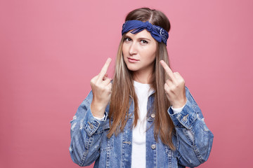 Irritated hippy female being fed up with relations with lover, keeps hands in fuck you sign, wears fashionable denim clothes in hippie style, isolated over pink background. Annoyance and subculture.
