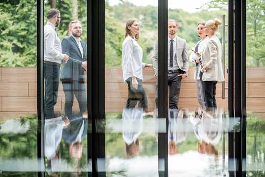 Business People Having A Break Standing Outdoors On The Office Balcony Overlooking On The Park. View Through The Window With Reflection