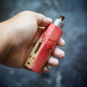 high end red natural stabilized wood box mods with rebuildable dripping atomizer vape gear in hand on dark grey texture background, vaporizer equipment, selective focus with shallow depth of field