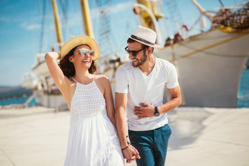 Happy young couple walking by the harbor of a touristic sea resort with sailboats on background