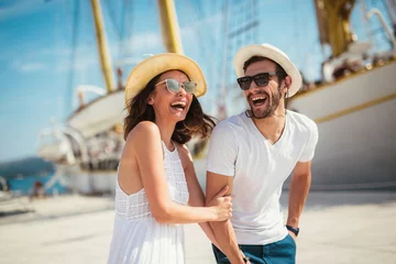 Fotobehang Happy young couple walking by the harbor of a touristic sea resort with sailboats on background © Mediteraneo