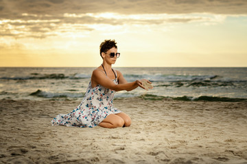 Fototapeta na wymiar beautiful woman in a dress sitting on the beach and scattering the sand at sunset