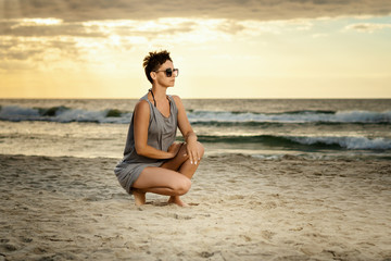 Fototapeta na wymiar beautiful woman in a gray dress crouches on the sand at the beach at sunset