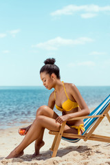 african american woman applying sunscreen lotion on skin while sitting on deck chair on sandy beach