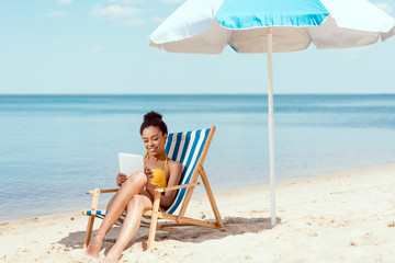 african american woman relaxing on deck chair and using digital tablet under beach umbrella in...