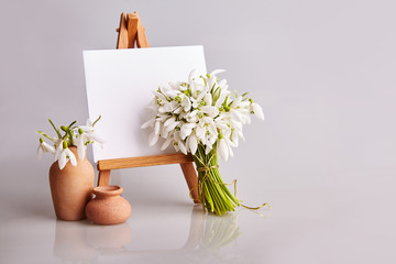 Bouquet of snowdrops and a small easel with a white paper and mini jars on a grey background. Greeting card with a space for text