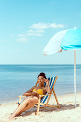 smiling african american woman relaxing on deck chair and listening music in earphones with...
