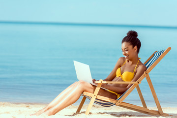 smiling african american female freelancer sitting on deck chair and using laptop on sandy beach