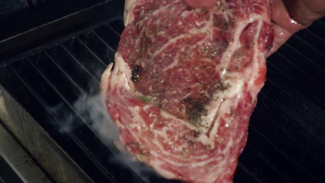 Cooking chief puts uncooked meat piece on hot smoking grill with fire underneath using metal forceps, Close up slow motion.