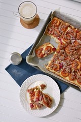 Top view of apricot and rhubarb pie with streusel on top in cake tin, pieces of pie on white plate on dark blue place mat, icing sugar tin, cup with tea with foam milk, placed on white table, sunny