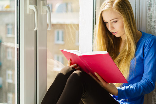 Young woman sitting at windowsill reading book